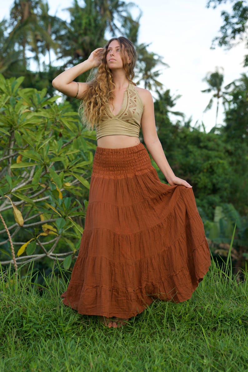 May include: A woman wearing a two-piece outfit outdoors. The top is a light green halter top with a geometric design. The skirt is a long, tiered, rust-colored skirt. The background features lush green foliage and palm trees.