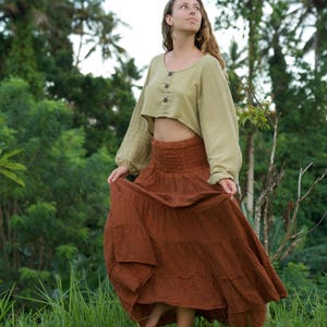 May include: A woman wearing a two-piece outfit in a natural setting. The outfit includes a light green, long-sleeved crop top with wooden buttons and a long, tiered, rust-colored skirt. The skirt is flowing, and the woman is standing in tall green grass.