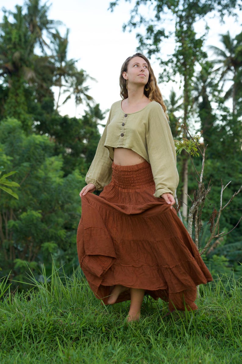 May include: A woman wearing a two-piece outfit in a natural setting. The outfit includes a light green, long-sleeved crop top with wooden buttons and a long, tiered, rust-colored skirt. The skirt is flowing, and the woman is standing in tall green grass.