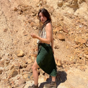 May include: A woman with long brown hair wearing a white tank top and a green wrap skirt. She is standing in a rocky, desert-like landscape.