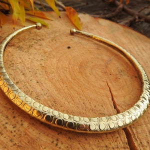 May include: A gold-toned brass choker necklace with a textured, circular pattern. The necklace is on a wooden surface.