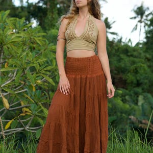 May include: A woman wearing a two-piece outfit outdoors. The top is a light olive green halter with a geometric design. The bottom is a long, tiered, rust-colored skirt. The background features lush green foliage and grass.