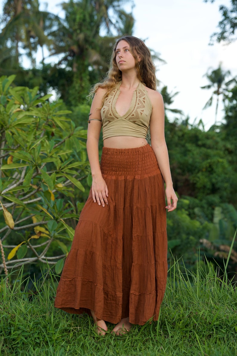 May include: A woman wearing a two-piece outfit outdoors. The top is a light olive green halter with a geometric design. The bottom is a long, tiered, rust-colored skirt. The background features lush green foliage and grass.