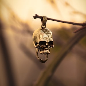 Peut inclure: Un pendentif en forme de crâne doré avec un design détaillé suspendu à une branche. Le crâne a une bouche grande ouverte et des dents visibles.