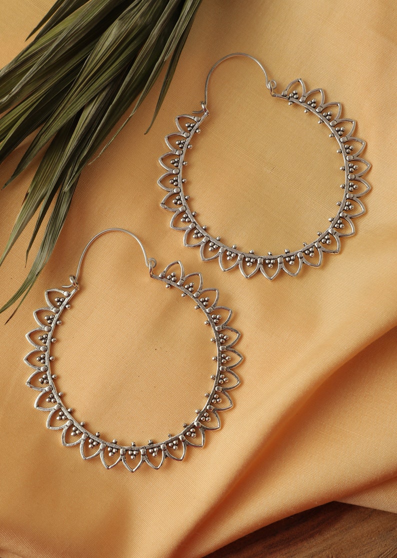 May include: A pair of silver hoop earrings with a delicate, intricate design. The earrings are circular and feature a scalloped edge with small, round accents.