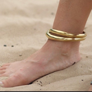 K&ouml;nnte beinhalten: Nahaufnahme eines Kn&ouml;chels, der zwei goldene Fu&szlig;kettchen tr&auml;gt. Die Fu&szlig;kettchen sind schlicht und aus dickem Metall gefertigt. Der Fu&szlig; der Person ruht auf Sand.