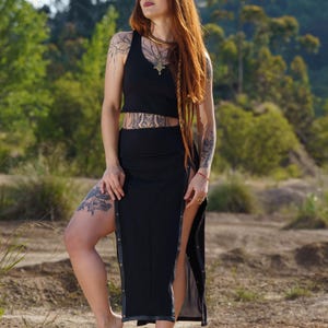Puede incluir: Un top corto negro y una falda larga a juego con una abertura alta. La falda tiene botones decorativos a lo largo de la abertura. La modelo tiene el pelo largo rojo y tatuajes. Se lleva un collar de oro.