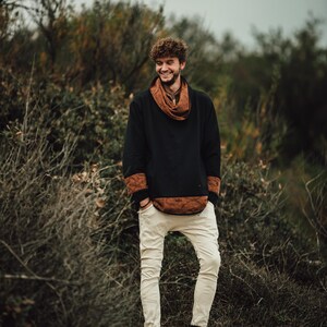 Puede incluir: Una persona con una camisa negra de manga larga con detalles marrones estampados y una bufanda a juego. La persona tambi&eacute;n lleva pantalones blancos rotos y botas marrones. El fondo presenta follaje verde y marr&oacute;n.