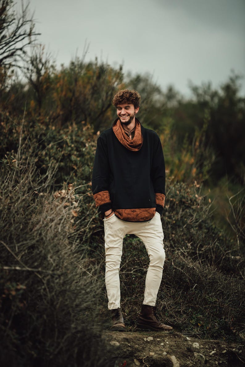 Puede incluir: Una persona con una camisa negra de manga larga con detalles marrones estampados y una bufanda a juego. La persona tambi&eacute;n lleva pantalones blancos rotos y botas marrones. El fondo presenta follaje verde y marr&oacute;n.