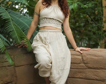 May include: A woman wearing a white halter top with a geometric pattern and matching white pants. She is standing in front of a stone wall with green foliage behind her.