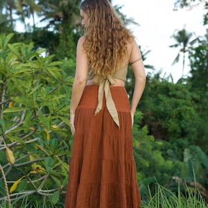 May include: A woman wearing a rust-colored tiered maxi skirt and a light green halter top with a bow in the back. The woman is standing in a grassy area with lush green foliage and palm trees in the background.