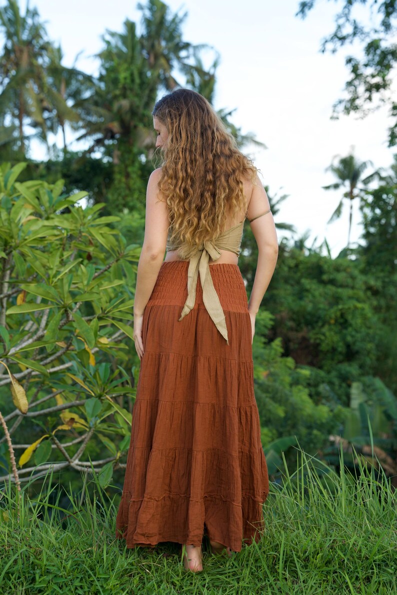 May include: A woman wearing a rust-colored tiered maxi skirt and a light green halter top with a bow in the back. The woman is standing in a grassy area with lush green foliage and palm trees in the background.