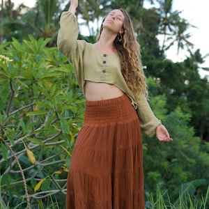 May include: A woman wearing a sage green long-sleeved crop top with three wooden buttons and a rust-colored tiered maxi skirt. The outfit is made of natural fabrics and is set against a lush green background.