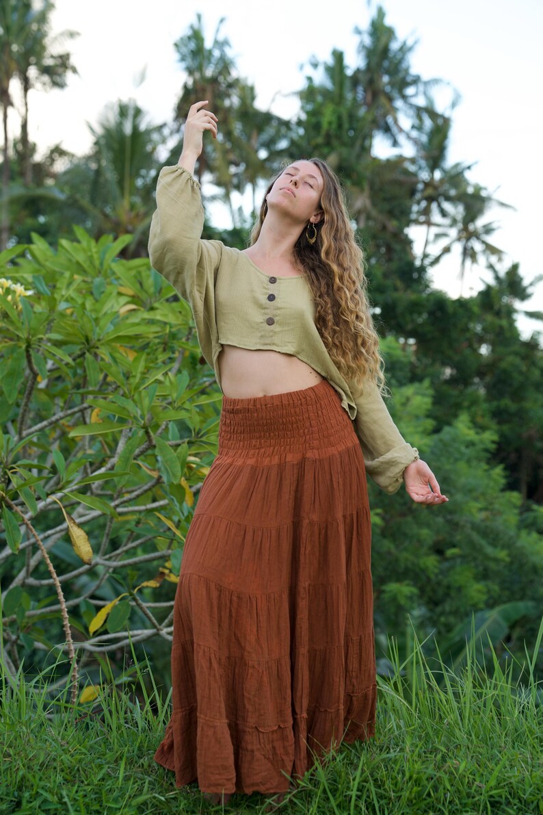 May include: A woman wearing a sage green long-sleeved crop top with three wooden buttons and a rust-colored tiered maxi skirt. The outfit is made of natural fabrics and is set against a lush green background.