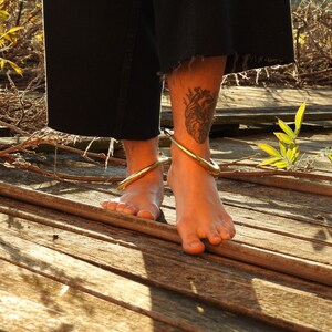 K&ouml;nnte beinhalten: Die nackten F&uuml;&szlig;e einer Person, die zwei goldene Fu&szlig;kettchen auf einer Holzoberfl&auml;che tr&auml;gt. Die Person hat ein Herz-Tattoo an ihrem Kn&ouml;chel.
