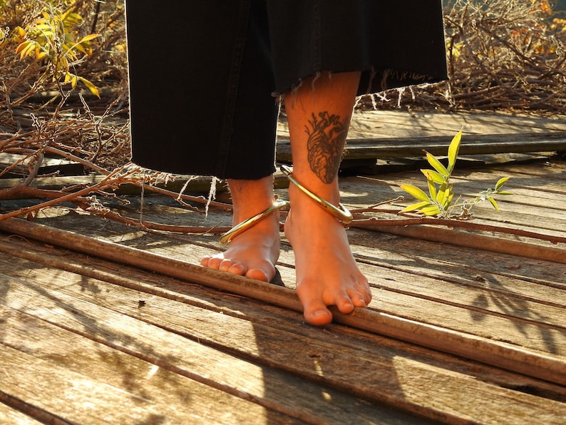 K&ouml;nnte beinhalten: Die nackten F&uuml;&szlig;e einer Person, die zwei goldene Fu&szlig;kettchen auf einer Holzoberfl&auml;che tr&auml;gt. Die Person hat ein Herz-Tattoo an ihrem Kn&ouml;chel.