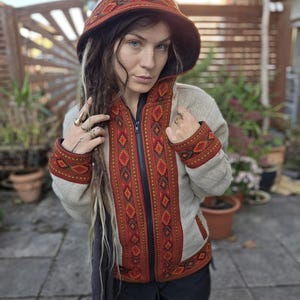 Könnte beinhalten: Kapuzenjacke mit geometrischem Muster in Rot, Orange und Braun. Die Jacke hat einen Reißverschluss und ist aus einem strukturierten, hellfarbigen Stoff gefertigt. Die Kapuze ist spitz und hat einen passenden gemusterten Besatz.