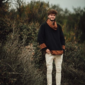 Puede incluir: Una camisa negra de manga larga con detalles marrones estampados en el cuello, los pu&ntilde;os y el dobladillo. El conjunto se combina con pantalones blancos rotos y botas marrones. La persona est&aacute; de pie al aire libre en un entorno natural.