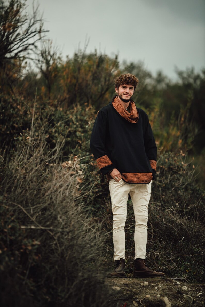 Puede incluir: Una camisa negra de manga larga con detalles marrones estampados en el cuello, los pu&ntilde;os y el dobladillo. El conjunto se combina con pantalones blancos rotos y botas marrones. La persona est&aacute; de pie al aire libre en un entorno natural.
