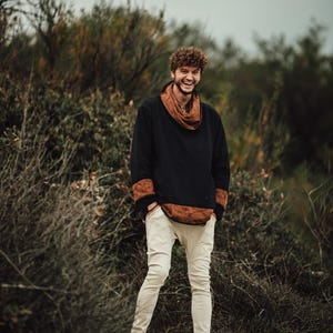 Puede incluir: Una persona con un su&eacute;ter negro y marr&oacute;n con una bufanda marr&oacute;n y pantalones blancos. La persona est&aacute; sonriendo y de pie afuera frente a unos arbustos. La persona lleva botas marrones.