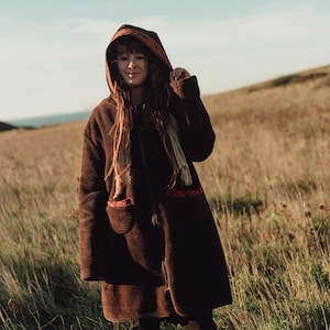 Può includere: Un lungo cappotto marrone con cappuccio, chiusura a zip e due tasche anteriori. Il cappotto ha un aspetto strutturato e viene indossato all'aperto in un campo di erba alta. Il cappuccio è alzato e il cappotto sembra essere fatto di un materiale caldo.