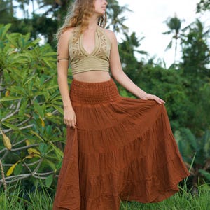 May include: A woman wearing a halter top and a tiered maxi skirt. The halter top is a light olive green color with a detailed pattern. The skirt is a rust color and has multiple tiers. The woman is standing in a grassy area.