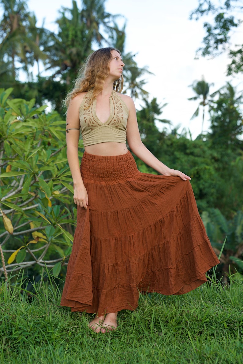 May include: A woman wearing a halter top and a tiered maxi skirt. The halter top is a light olive green color with a detailed pattern. The skirt is a rust color and has multiple tiers. The woman is standing in a grassy area.