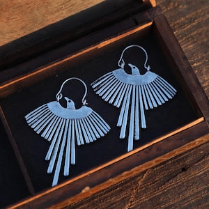 May include: A pair of silver-colored bird-shaped earrings displayed inside a wooden box. The earrings feature a bird design with outstretched wings and long, vertical lines. The box has a dark interior and a natural wood exterior.
