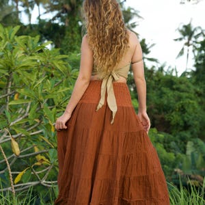 May include: A woman wearing a rust-colored tiered maxi skirt and a light olive green halter top with a bow in the back. The skirt is made of a textured fabric. The background features lush green foliage and palm trees.