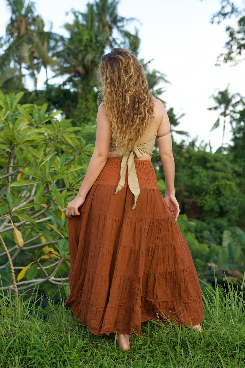 May include: A woman wearing a rust-colored tiered maxi skirt and a light olive green halter top with a bow in the back. The skirt is made of a textured fabric. The background features lush green foliage and palm trees.