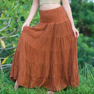 May include: A tiered, rust-colored maxi skirt with a smocked waistband. The skirt is paired with a halter top in a light olive green color. The outfit is worn outdoors in a grassy area.
