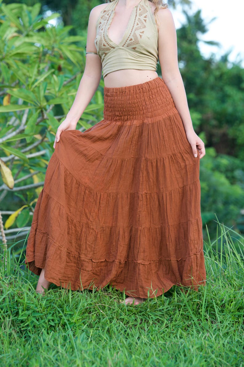 May include: A tiered, rust-colored maxi skirt with a smocked waistband. The skirt is paired with a halter top in a light olive green color. The outfit is worn outdoors in a grassy area.