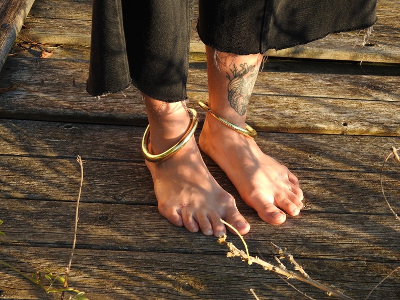 K&ouml;nnte beinhalten: Zwei goldene Fu&szlig;kettchen an nackten F&uuml;&szlig;en. Die Fu&szlig;kettchen sind dick und rund und werden an den Kn&ouml;cheln einer Person getragen, die auf einer Holzoberfl&auml;che steht.