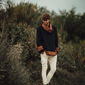 Puede incluir: Una persona con una camisa negra de manga larga con detalles marrones estampados y una bufanda a juego. La persona tambi&eacute;n lleva pantalones blancos y botas marrones. El fondo presenta follaje verde y marr&oacute;n bajo un cielo nublado.