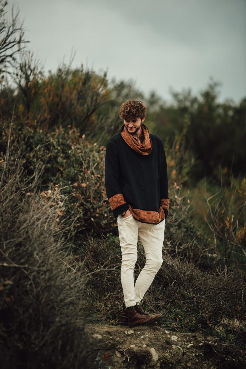 Puede incluir: Una persona con una camisa negra de manga larga con detalles marrones estampados y una bufanda a juego. La persona tambi&eacute;n lleva pantalones blancos y botas marrones. El fondo presenta follaje verde y marr&oacute;n bajo un cielo nublado.
