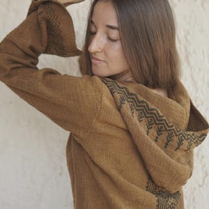 May include: A brown hooded long-sleeve top featuring a textured fabric and a dark brown geometric pattern along the hood and sleeve edges. The garment has a relaxed fit with wide sleeves and a hood that rests against the back of the shoulders.