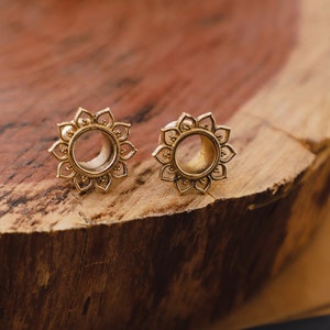 May include: Two gold-toned metal ear gauges with intricate floral designs. The gauges are circular with a hollow center and feature a detailed pattern of small circles and petals.