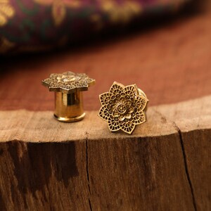 May include: Two gold-colored metal ear gauges with a detailed floral design. The gauges are on a wooden surface.