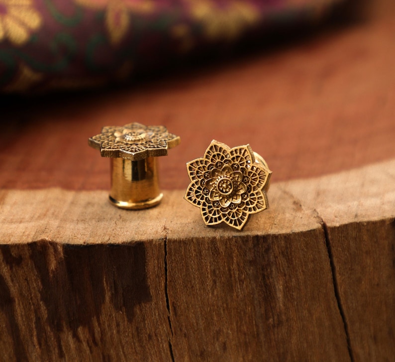 May include: Two gold-colored metal ear gauges with a detailed floral design. The gauges are on a wooden surface.