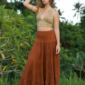 May include: A woman wearing a halter top and a long, tiered skirt. The halter top is a light olive green with a detailed neckline. The skirt is a rust color and reaches the ground. The background is lush green foliage.