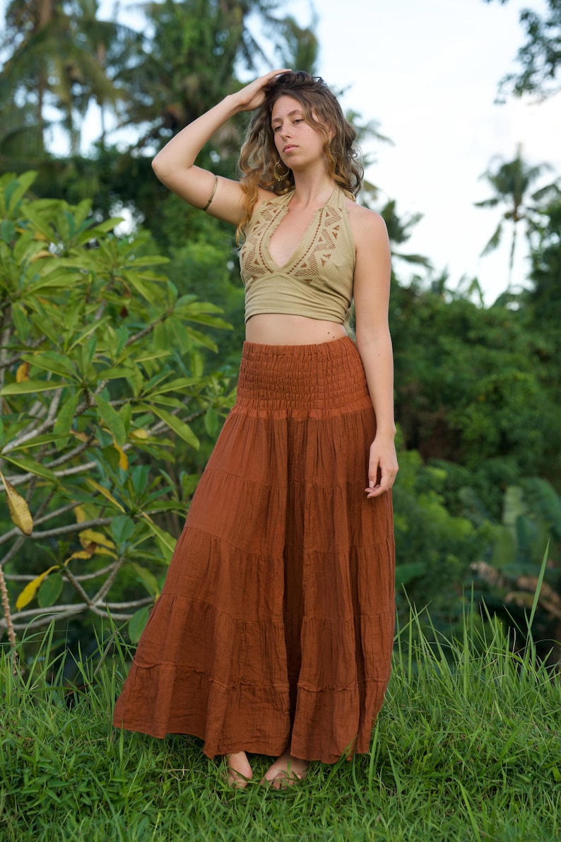 May include: A woman wearing a halter top and a long, tiered skirt. The halter top is a light olive green with a detailed neckline. The skirt is a rust color and reaches the ground. The background is lush green foliage.