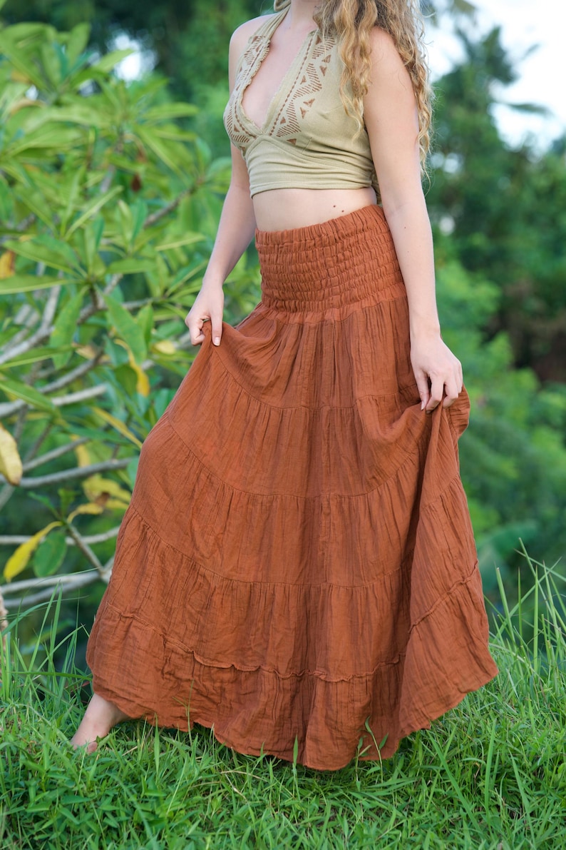 May include: A rust-colored, tiered maxi skirt with a smocked waistband. The skirt is paired with a halter top in a light olive green color. The outfit is suitable for casual wear and is made of a lightweight fabric.