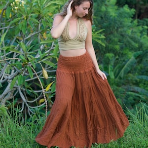 May include: A woman wearing a halter top and a long, tiered skirt. The halter top is a light olive green with a patterned design. The skirt is a rust color and has a gathered waistband. The background is lush green foliage.