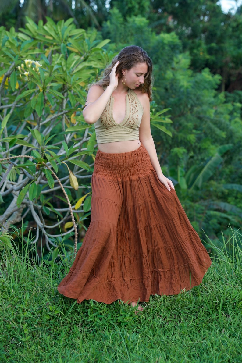 May include: A woman wearing a halter top and a long, tiered skirt. The halter top is a light olive green with a patterned design. The skirt is a rust color and has a gathered waistband. The background is lush green foliage.
