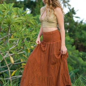 May include: A woman wearing a light green halter top and a long, tiered, rust-colored skirt. The skirt has a gathered waistband and flows down to the ground. The background features lush green foliage.