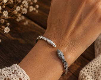 Puede incluir: Un brazalete tipo puño de tono plateado que presenta dos cabezas de serpiente detalladas frente a frente en la abertura. La superficie metálica tiene un acabado oxidado que resalta las escamas texturizadas y los ojos de las serpientes.