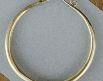May include: A gold-colored, circular necklace with a simple design. The necklace has a smooth, polished surface and features a hook-and-eye closure at the top. The jewelry is displayed on a gray surface with a wooden background.