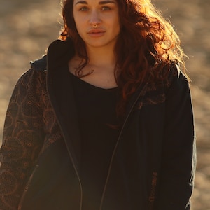 May include: A woman with long, red hair is wearing a black zip-up hoodie with a brown patterned design on the sleeves. The hoodie has a hood and a zipper closure.