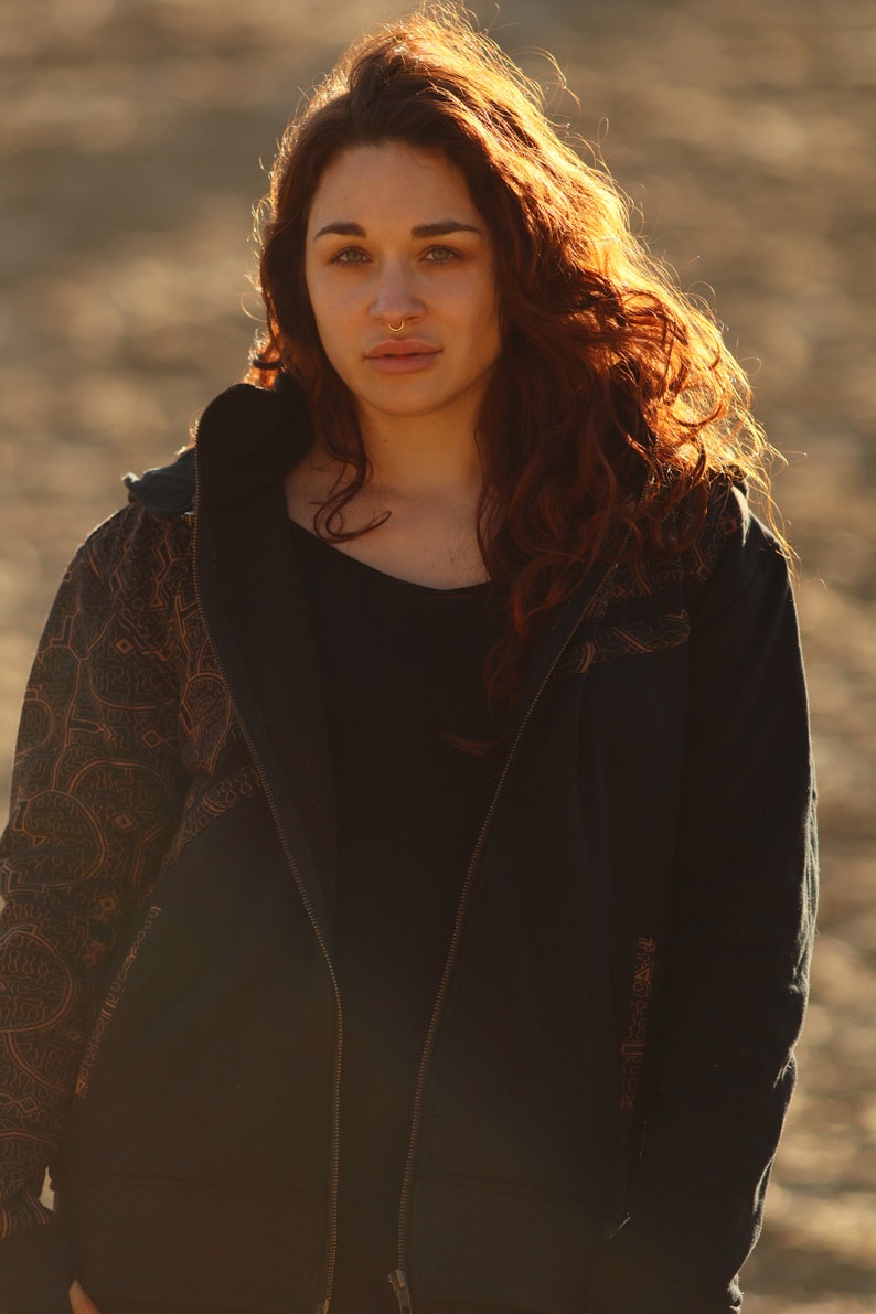 May include: A woman with long, red hair is wearing a black zip-up hoodie with a brown patterned design on the sleeves. The hoodie has a hood and a zipper closure.