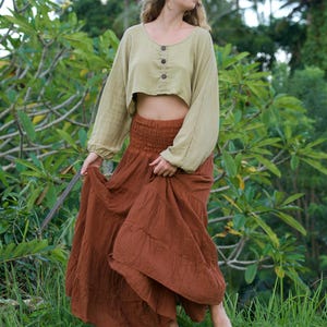 May include: A woman wearing a two-piece outfit. The top is a light green, long-sleeved, cropped blouse with three buttons. The bottom is a long, flowing, rust-colored skirt. The outfit is worn outdoors in a natural setting.